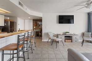 A modern kitchen with a bar area, a dining space with chairs, a seating area with a sofa, and a television mounted on the wall. - Salt Ponds | Work & Relax in Key West | Poolside Condo • Balcony • Near Beach (Key west)