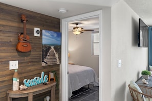 A room with a wooden wall, a guitar, a bed, a ceiling fan, and a TV mounted on the wall. - Salt Ponds | Work & Relax in Key West | Poolside Condo • Balcony • Near Beach (Key west)