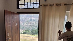 A bedroom with a large window, a wooden door, and a person standing by the window.