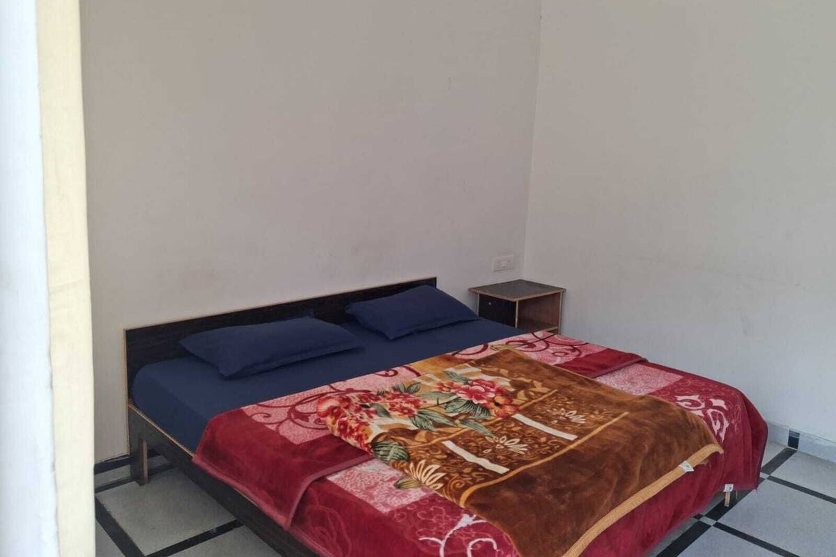 Uma cama de solteiro com colcha florida e duas almofadas, uma mesinha de cabeceira pequena e uma parede lisa.