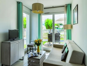 Una sala moderna con un sofá, un televisor sobre un mueble, una mesita con un florero y vistas al exterior.