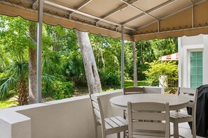 Eine überdachte Terrasse im Freien mit einem runden Tisch und Stühlen, umgeben von üppiger Bepflanzung und Bäumen.