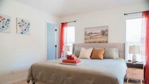 Una camera da letto con un letto, un comodino, una lampada, una porta e due finestre con tende.