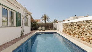 Una piscina con un muro de piedra a un lado y un edificio blanco con contraventanas verdes al otro.