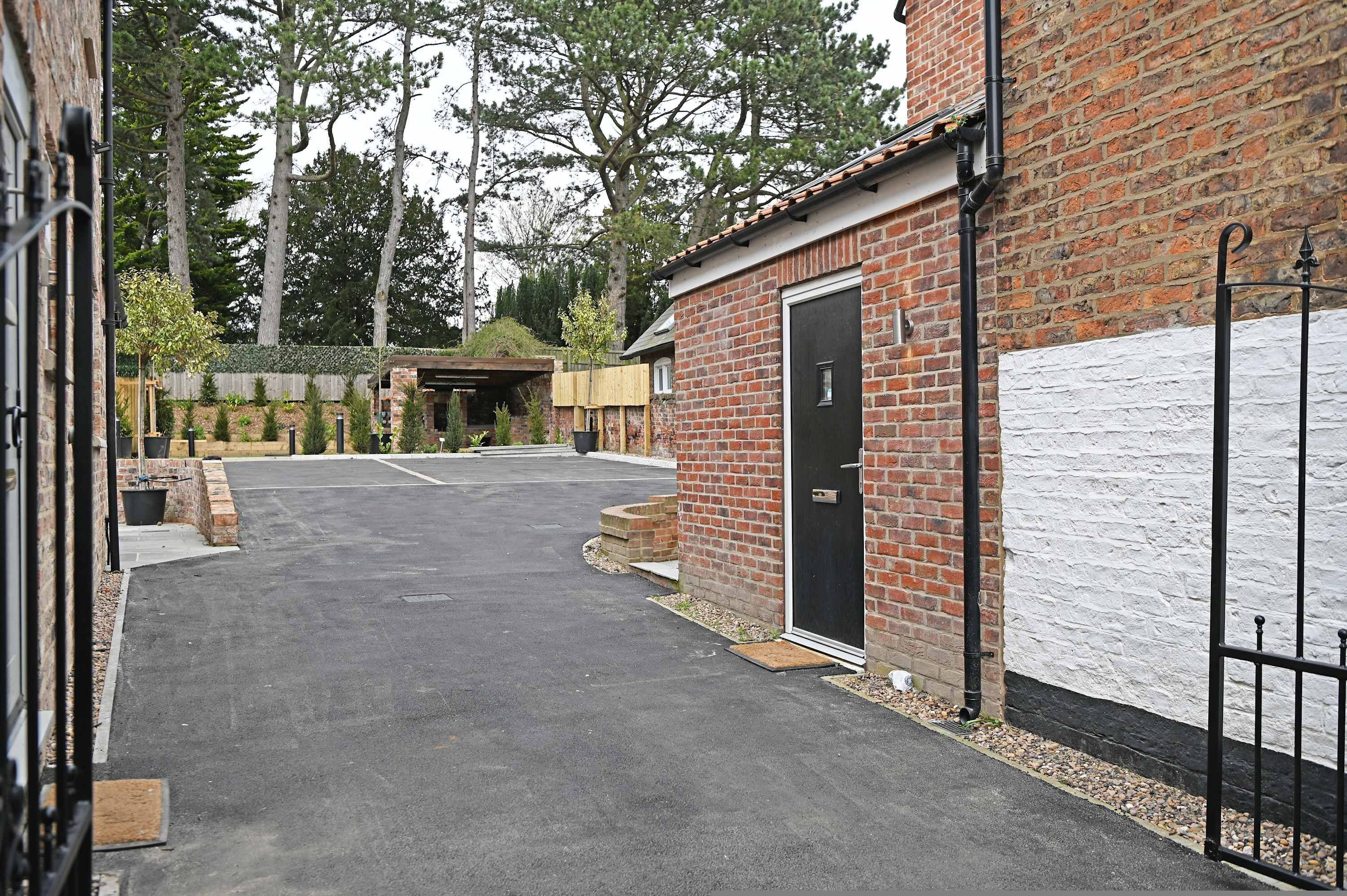 A paved driveway leading to a brick building with a black door, surrounded by trees and a white wall.
