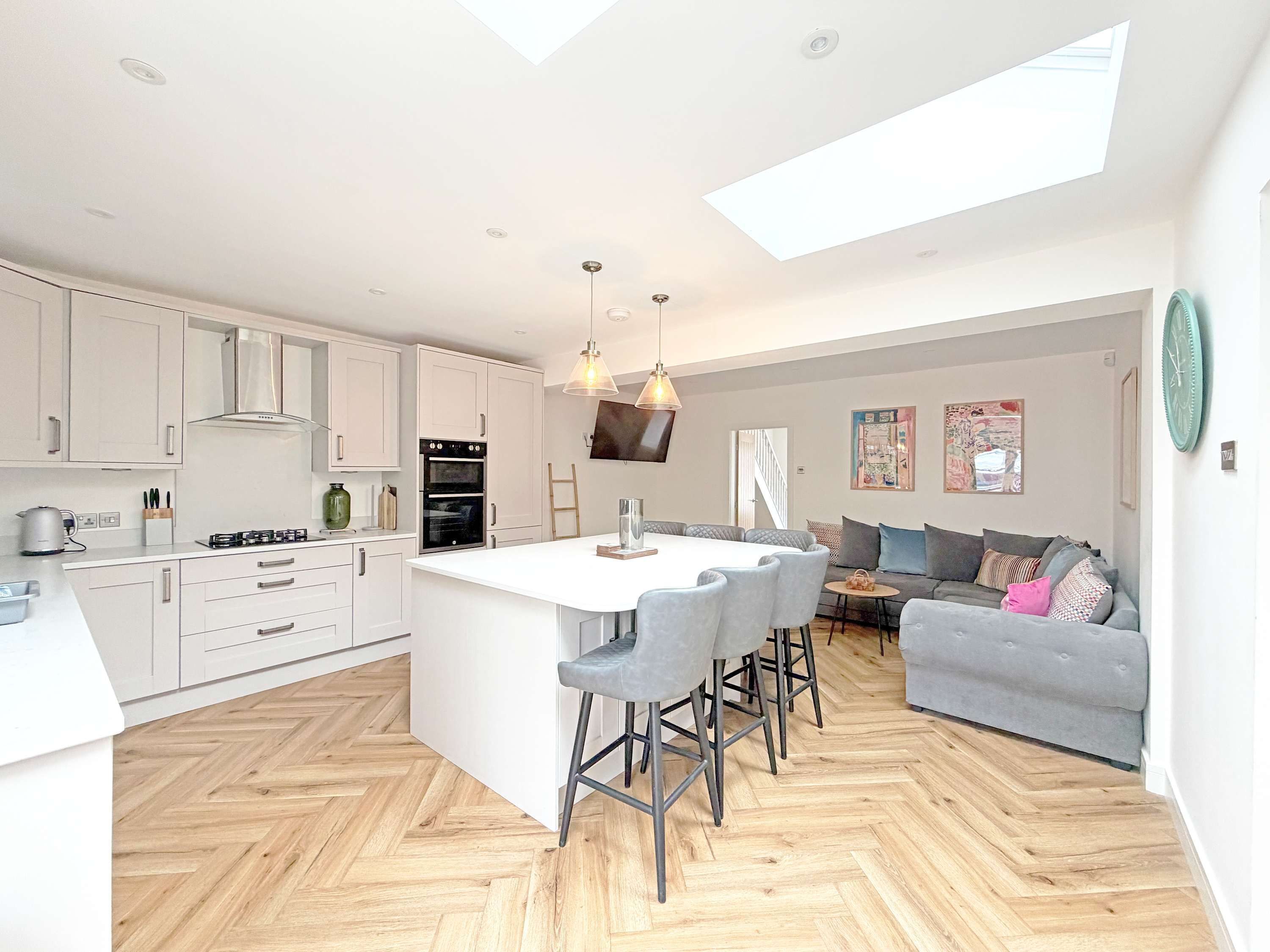 A modern kitchen with a central island, a dining area with bar stools, a grey sofa, and a herringbone wooden floor.
