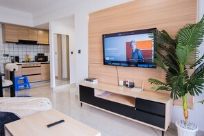 Ein modernes Wohnzimmer mit Flachbildfernseher an einer Holzwand, einer Couch, einem Couchtisch, einem blauen Hocker, einem Blumentopf und einer Küche im Hintergrund.