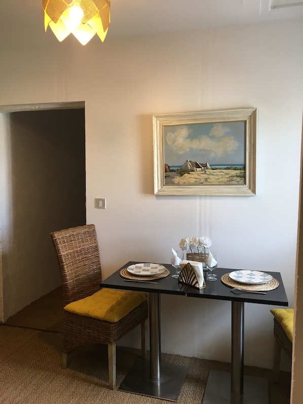 A dining area with a table set for two, a wicker chair, a painting on the wall, and a hanging light fixture. - Dwelling in farmhouse with independent entrance (no shared rooms) (Pertuis)