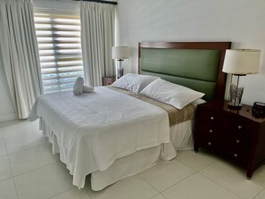 Un lit bien fait, avec du linge de lit blanc, une tête de lit recouverte d’un tissu vert et deux lampes de chevet.