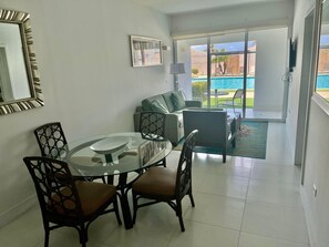 A modern living room with a glass dining table, a sofa, and a view of a pool through large sliding glass doors. - Arubaslife  Palm Beach Aruba · 2BR/2BA  6 Pax max (Noord)