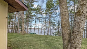 A building with a sloped roof, a grassy yard, and a lake with trees in the background. - Leonards Cabin on Round Lake | Private Dock, 140ft Shoreline, Close to Town - New Rental (Hayward)