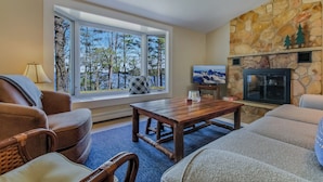 A living room with a stone fireplace, a large window with a view of trees, and a television. - Leonards Cabin on Round Lake | Private Dock, 140ft Shoreline, Close to Town - New Rental (Hayward)
