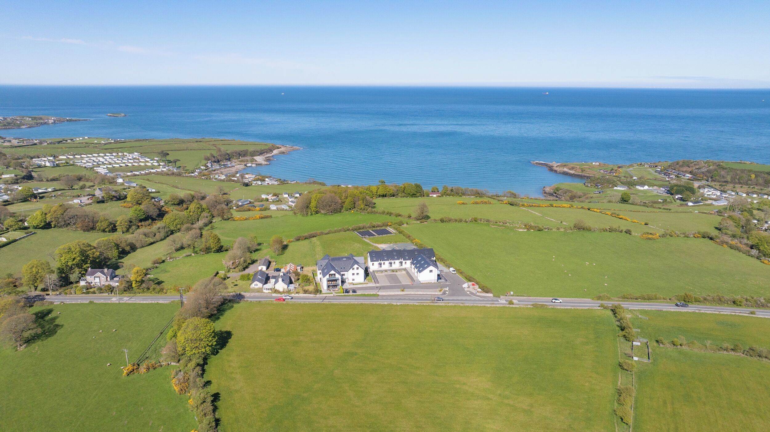 An aerial view of a coastal property with a large house surrounded by green fields, a road, and a body of water.