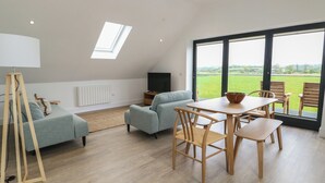 A modern living room with a sofa, a dining table with chairs, a TV, and a large window with a view of the outdoors. - Glynllifon 7 (Marianglas)