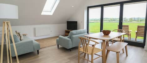 A modern living room with a sofa, a dining table with chairs, a TV, and a large window with a view of the outdoors.