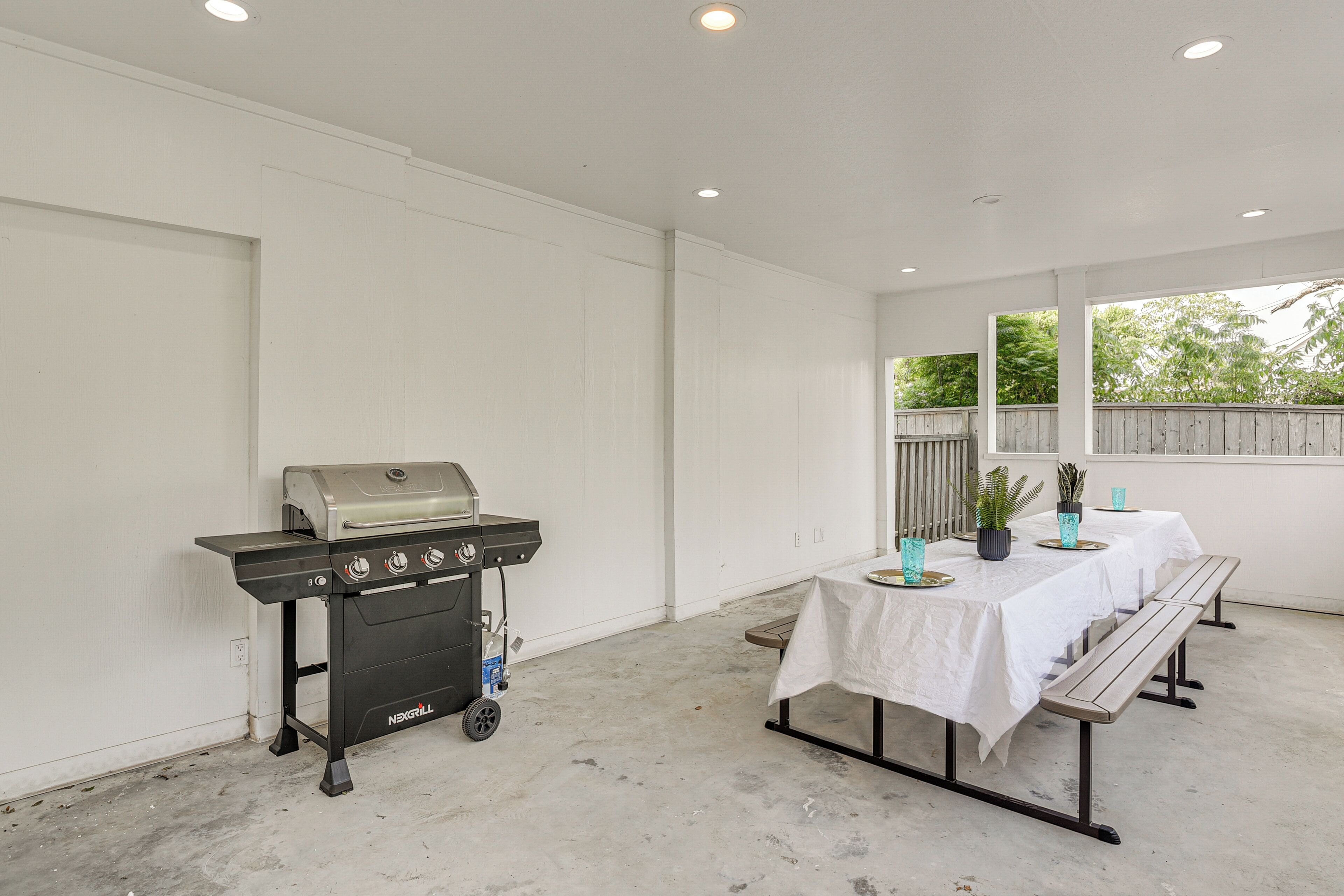 Ein überdachter Patio-Bereich mit Grill, Tischen mit weißen Tischdecken, Bänken und Blick auf Bäume durch große Fenster.