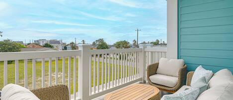 A porch with wicker furniture, a wooden table, and a view of a grassy area and buildings.