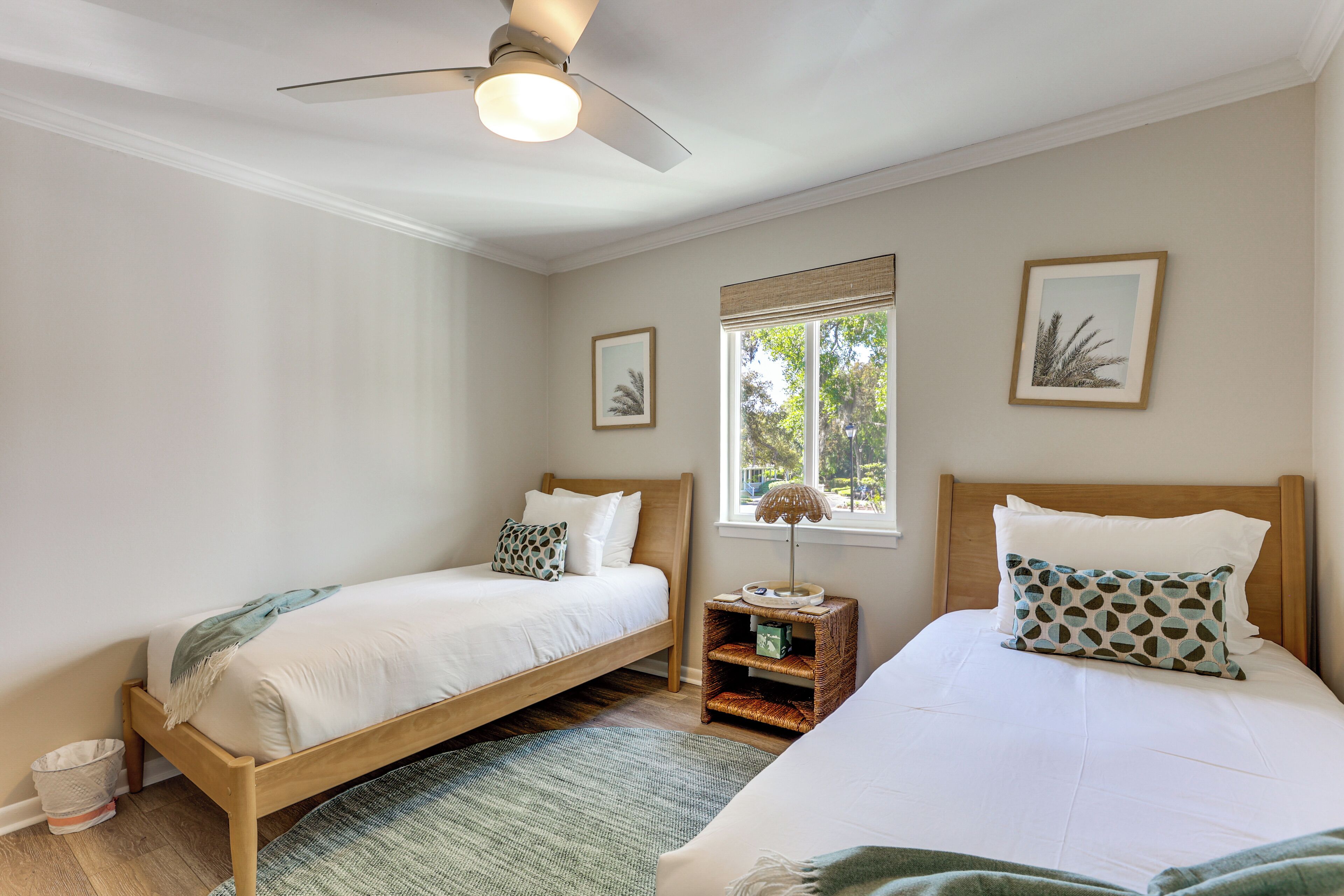 A bedroom with two single beds, a ceiling fan, a window with a view, and a small bedside table with a lamp.