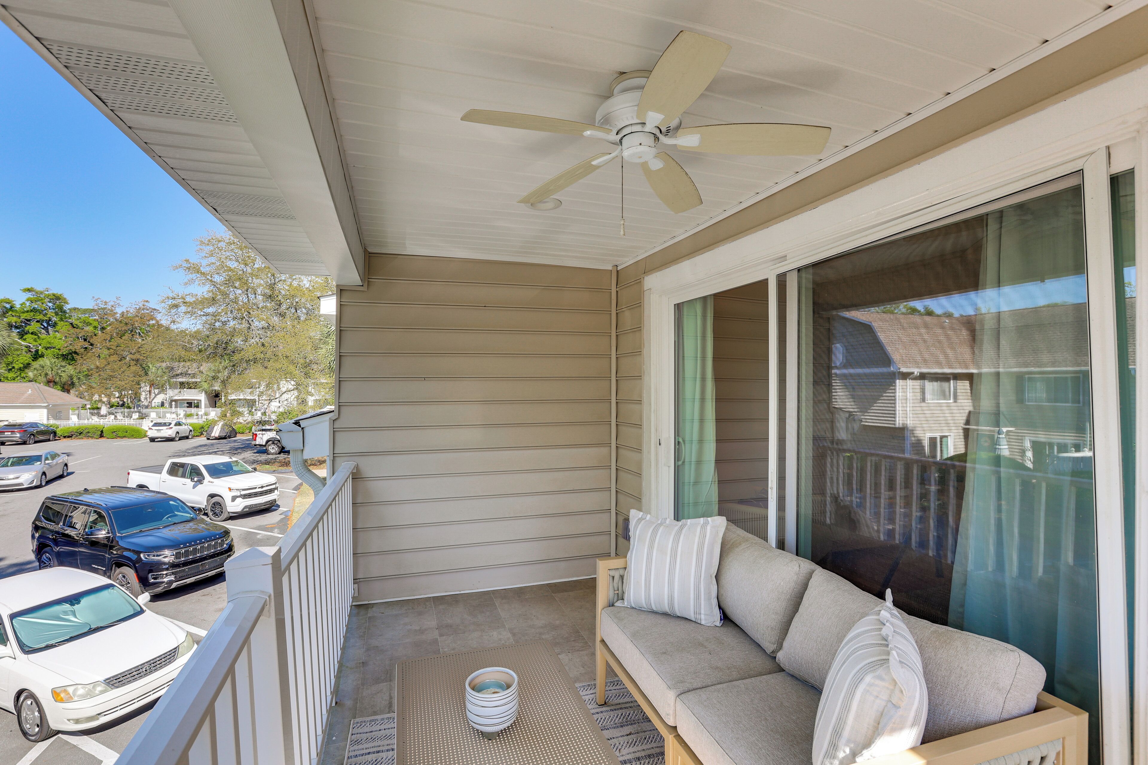 A balcony with a ceiling fan, a sofa, a table with a bowl, and a view of parked cars and houses.