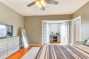 A bedroom with a bed, a TV, a ceiling fan, and a view of the living room through an open door.