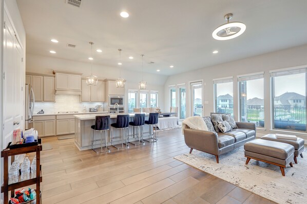 A spacious living area with a kitchen, bar stools, a sofa, and a large window offering a view. - 10 Min From Austin! High-end Home w/ Nice Backyard (Manor)