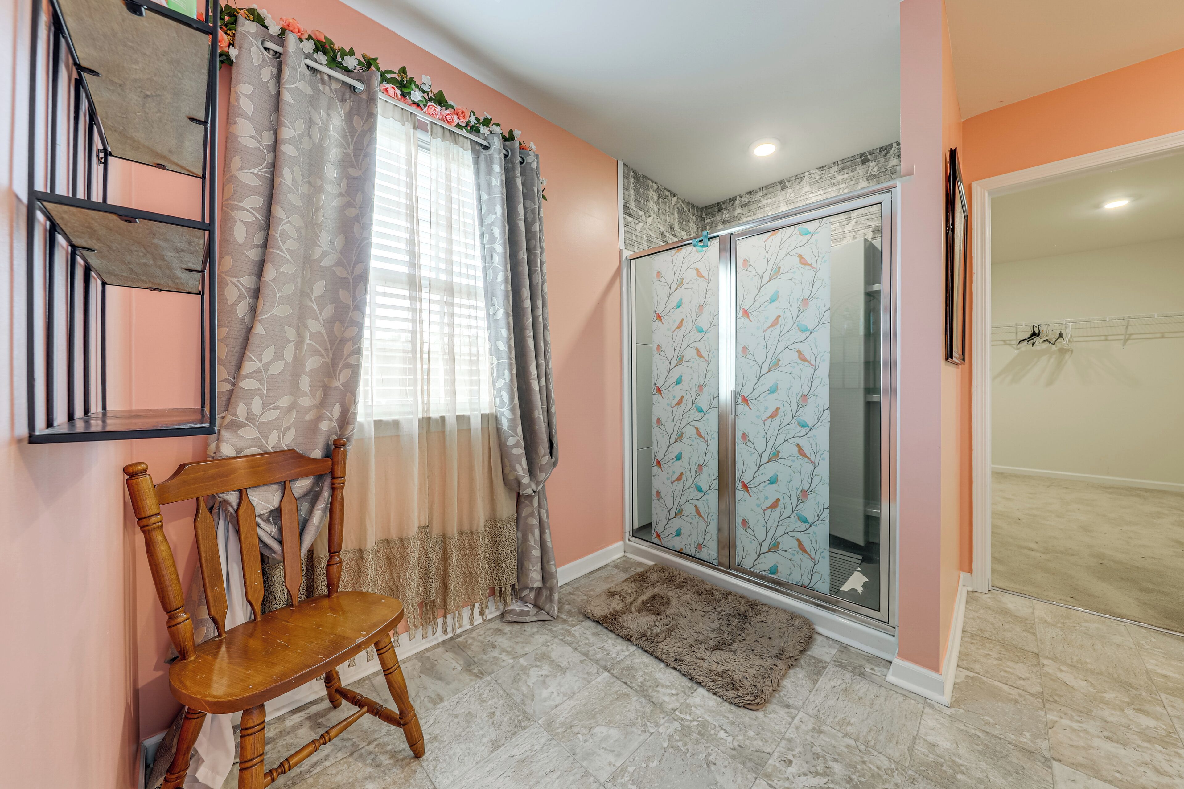 Une pièce avec une chaise en bois, une douche en verre ornée de motifs floraux, un rideau gris et un sol carrelé.