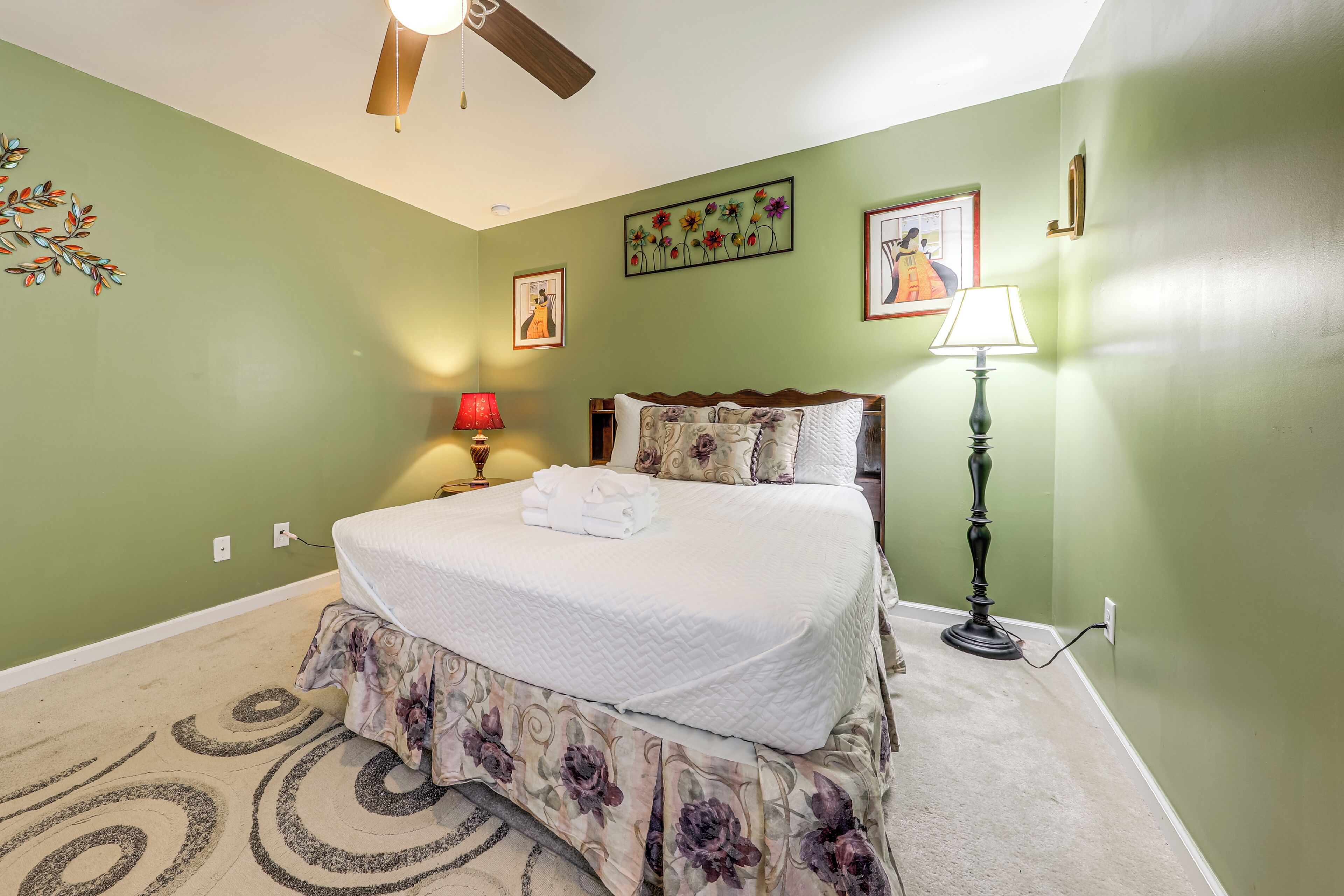 A bedroom with a bed, two bedside lamps, a ceiling fan, and a wall with floral artwork.