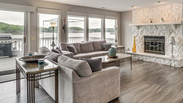Ein Wohnzimmer mit Sofa, Couchtisch und Kamin, großen Fenstern mit Blick auf ein Gewässer und einem Balkon mit Gartenmöbeln.