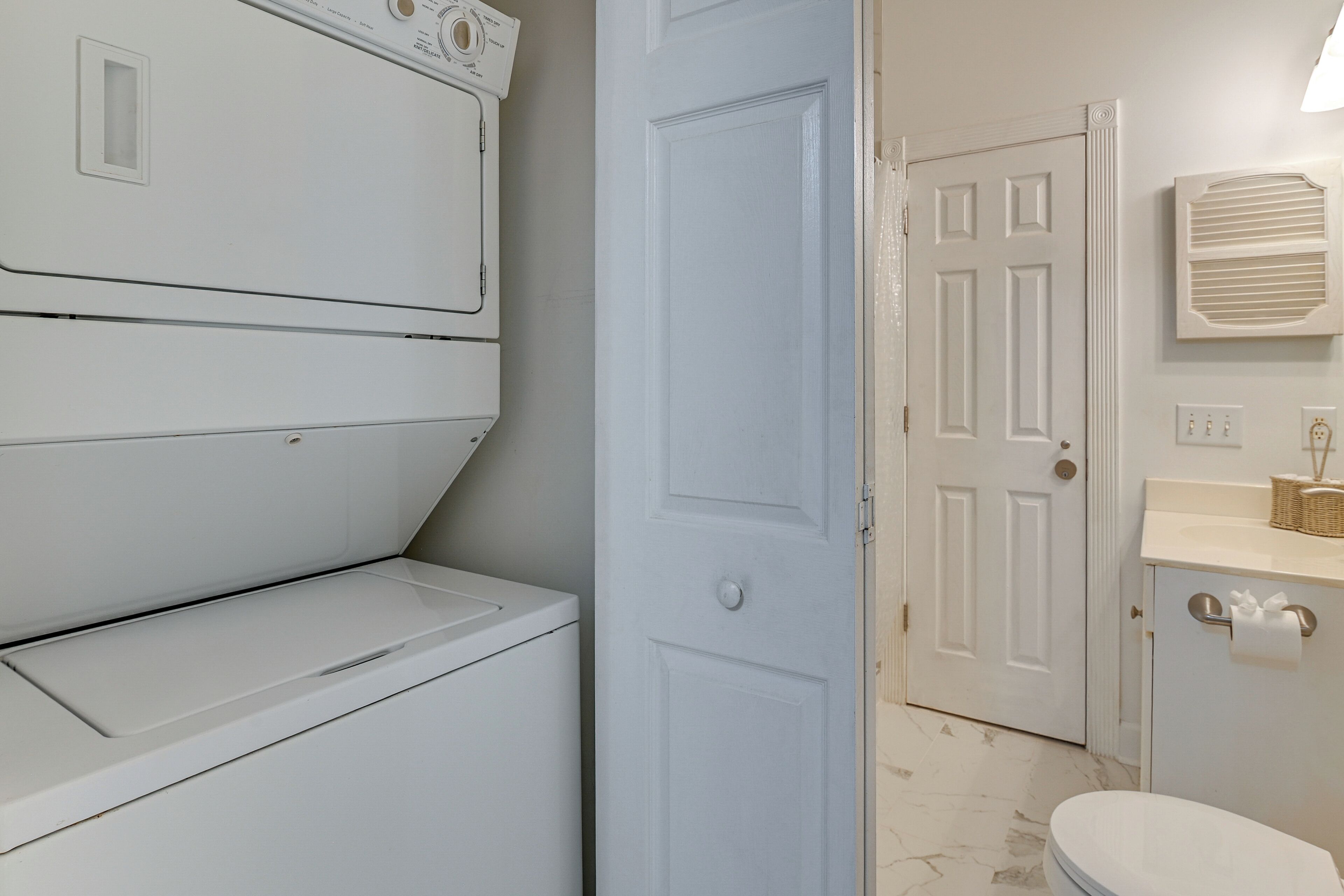 A laundry room with stacked washers and dryers, a white door, and a bathroom with a toilet, sink, and towel rack.