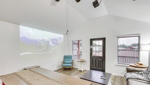 Una sala moderna con una pantalla de proyección grande, una mesa de madera, una silla azul, un sofá y una puerta con ventanal.