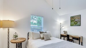 Un dormitorio con cama, mesita de noche, lámpara, ventana, escritorio, silla y un cuadro en la pared.
