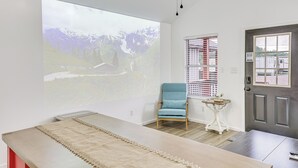 Una habitación con una pantalla de proyección que muestra un paisaje, un sillón azul, una mesita blanca y una puerta oscura.