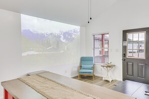 Una habitación con una pantalla de proyección que muestra un paisaje, un sillón azul, una mesita blanca y una puerta oscura.