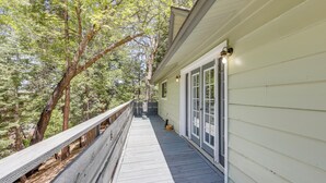 A wooden deck with a railing, a building with a white exterior, and trees in the background. - Gateway to Big Bear! High-country Home w/ Deck (Running Springs)