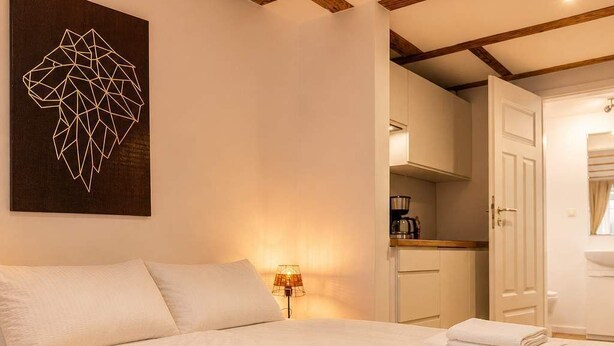 Ein Bett mit weißer Bettwäsche, ein geometrisches Wandbild, eine Lampe und ein durchgehender Blick auf einen Küchenbereich.