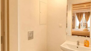 Ein kleines, sauberes Badezimmer mit Toilette, Waschbecken mit Spiegel und einem Fenster mit Vorhängen.