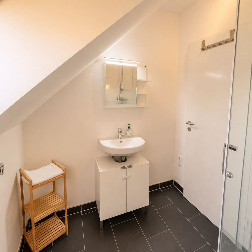 A bathroom with a white sink, mirror, and cabinet, a wooden shelf, and a glass shower door.