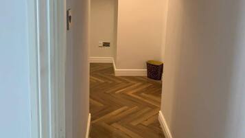 A hallway with a wooden herringbone floor, white walls, a ceiling light, and a small trash bin.