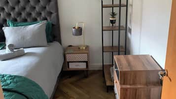 A bedroom with a tufted headboard, a bed with green bedding, a bedside table with a lamp, a wooden dresser, and a shelving unit with a plant.