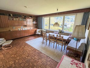 Ein gemütliches Wohnzimmer mit Holzboden, einem Esstisch mit Stühlen, einer Couch, einem Bücherregal und einem Fenster mit Blick auf Grünflächen.