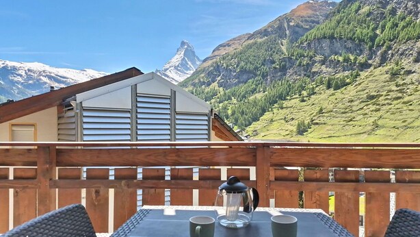 Eine Holzterrasse mit Tisch, Stühlen und einer Kaffeekanne, mit Blick auf einen Berg und eine Chalet im Hintergrund.