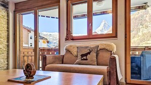 Ein gemütliches Zimmer mit Holztisch, einer Couch mit Felldecke und großen Fenstern mit Blick auf die Berge.
