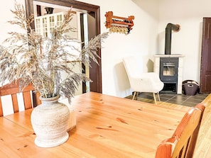 Ein Holzesstisch mit einer Vase gefüllt mit getrockneten Pflanzen, ein weißer Stuhl, eine Holzbank, ein Kamin und eine Wandregal mit Tassen.