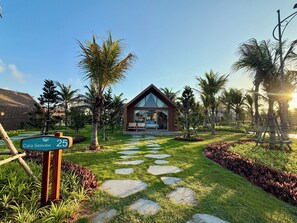 A modern house with a glass facade, surrounded by a well-maintained garden with palm trees and a stone pathway leading to the entrance.