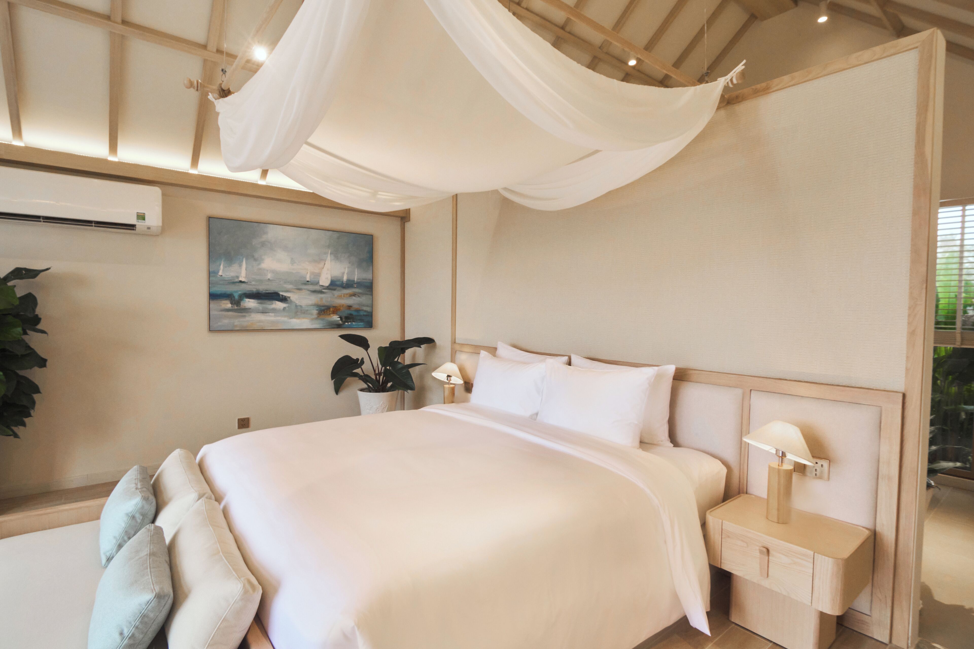 A bedroom with a large bed, bedside tables with lamps, a painting on the wall, and a potted plant.
