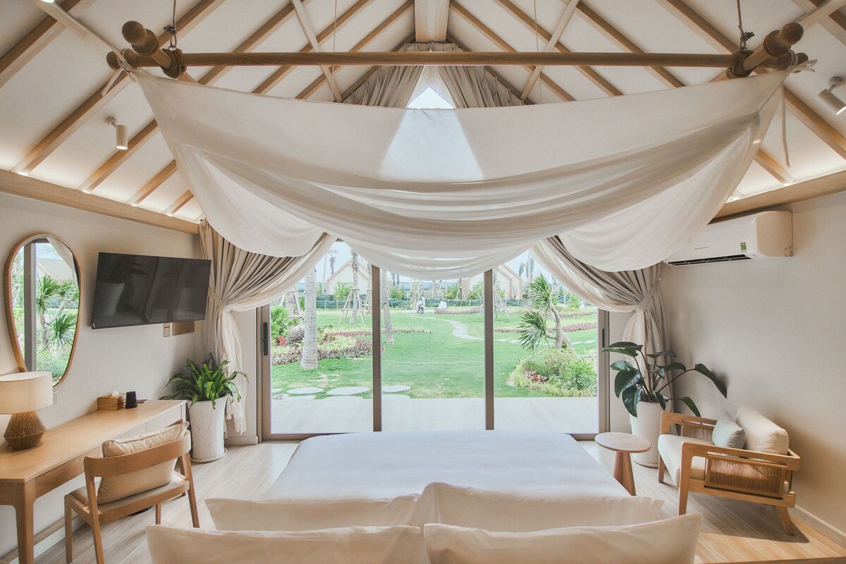 Quarto com uma cama grande, uma escrivaninha, uma cadeira, uma televisão e vista para um jardim.