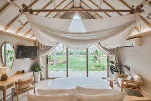 A bedroom with a large bed, a desk, a chair, a TV, and a view of a garden. - Carabeach Retreat (Cam Ranh)