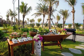 An outdoor dining area with a table set for a meal, surrounded by palm trees and a view of the sea.
