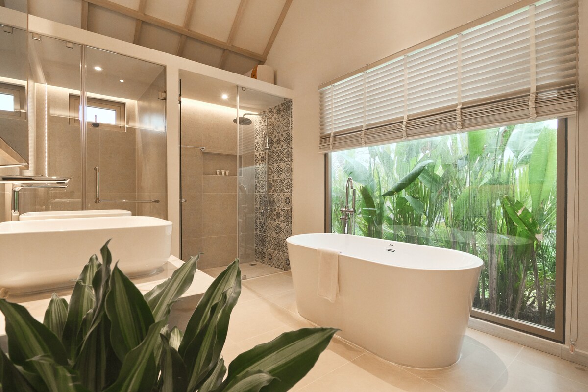 Un baño moderno con una bañera independiente, ducha con mampara de vidrio y un amplio ventanal con vistas al verdor.