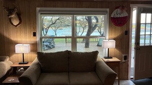 Ein gemütliches Zimmer mit einer Holzwand, einem großen Fenster mit Blick auf Bäume und einen Steg, einem Sofa, zwei Lampen und einer Tür mit einem Schild.